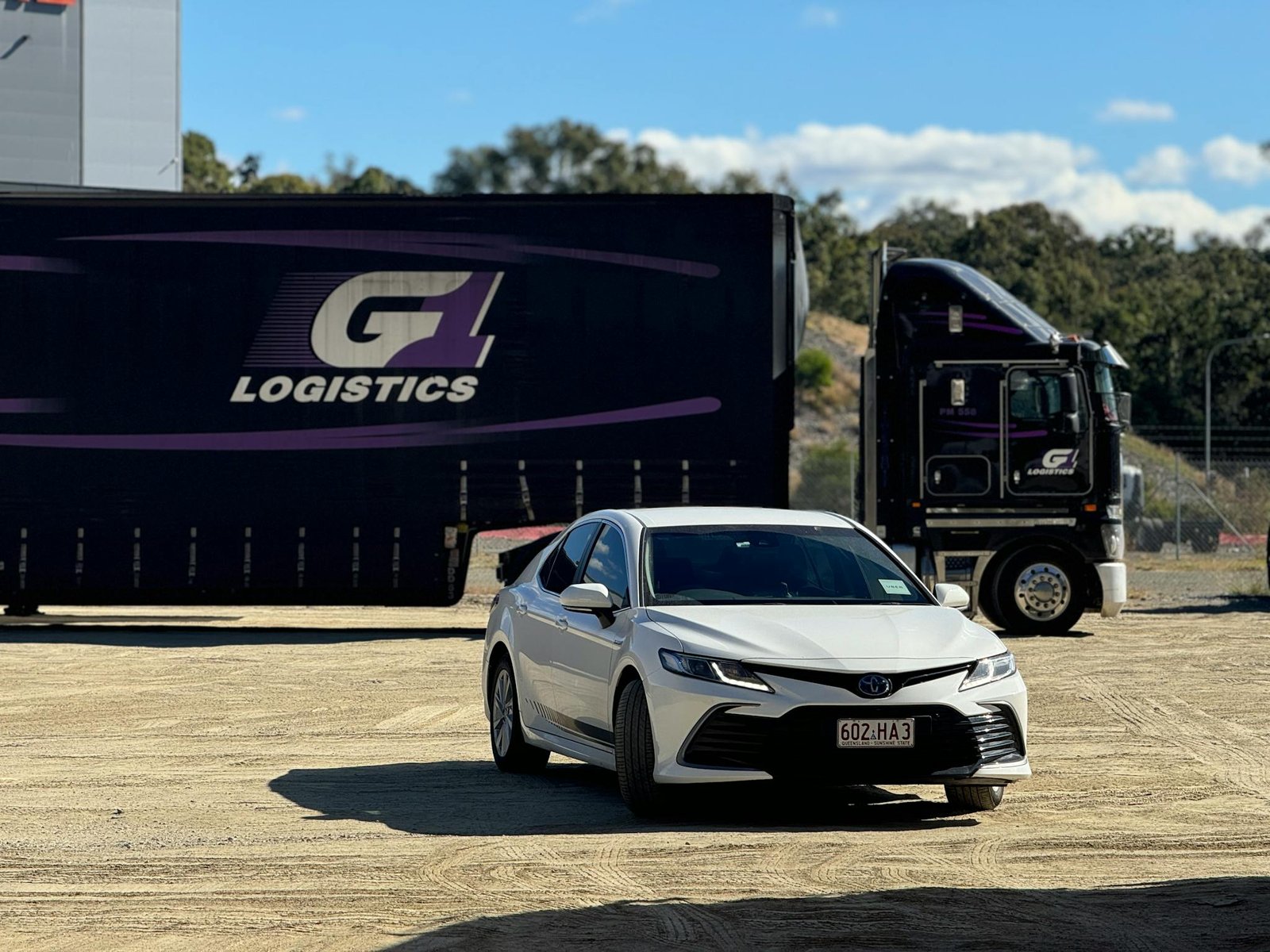 White sedan parked in front of a logistics truck at an outdoor loading area on a sunny day.
